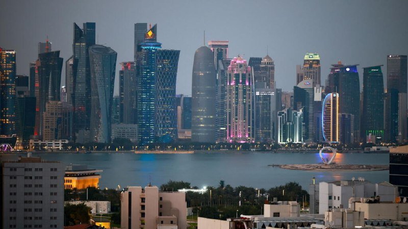 A modern waterfront city skyline with illuminated skyscrapers, including a distinctive building with a crosshatched facade, rises across a calm bay at dusk.