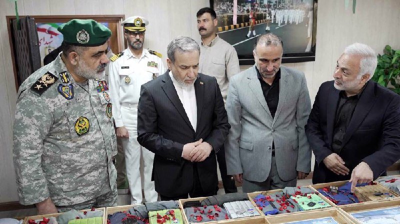 Six men, including one in military camouflage and another in a white naval uniform, examine displayed mechanical components laid out on tables in an indoor room.