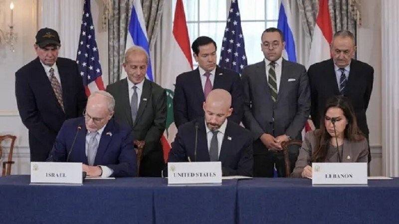 Officials sign documents at a table labeled "Israel," "United States," and "Lebanon," with five officials standing behind them flanked by U.S., Israeli, and Lebanese flags.