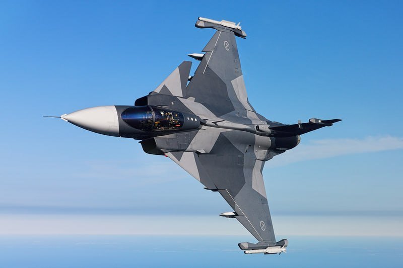 A gray camouflage fighter jet banks sharply against a clear blue sky, with a pilot visible in the cockpit.
