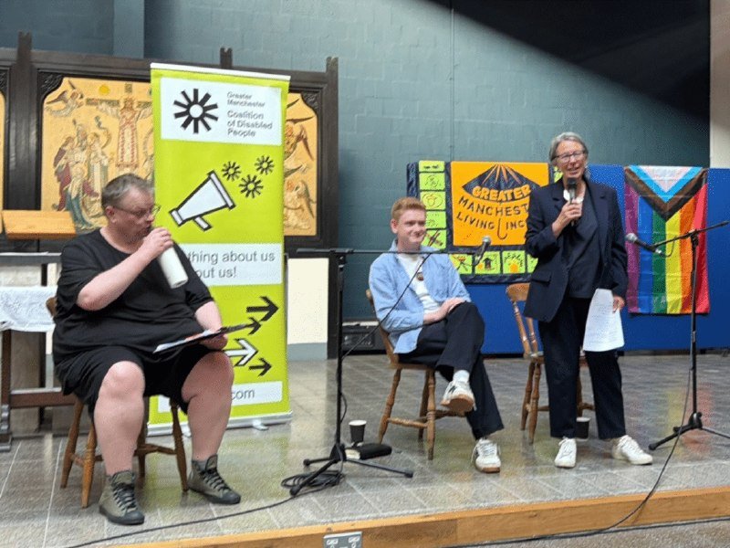 Three people sit on a stage with microphones, a green banner reading "Greater Manchester Coalition of Disabled People," and a Progress Pride flag visible in the background.