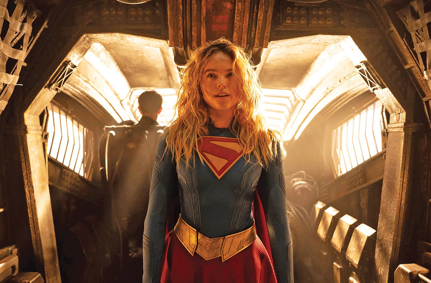 A blonde woman in a blue, red, and gold superhero costume stands in a dimly lit, industrial-looking corridor with arched metal framing and warm backlighting.