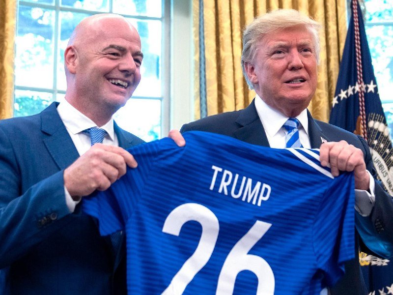 Two men in suits smile while holding up a blue soccer jersey reading "TRUMP 26" in the Oval Office, with a presidential flag visible behind them.