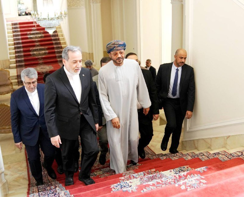 A group of men in business suits and traditional Middle Eastern attire walk together down a grand staircase covered with a red patterned carpet beneath a crystal chandelier.
