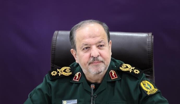 A man with a gray beard wearing a dark green military uniform with gold insignia sits in a black leather chair against a purple backdrop, facing forward near a microphone.