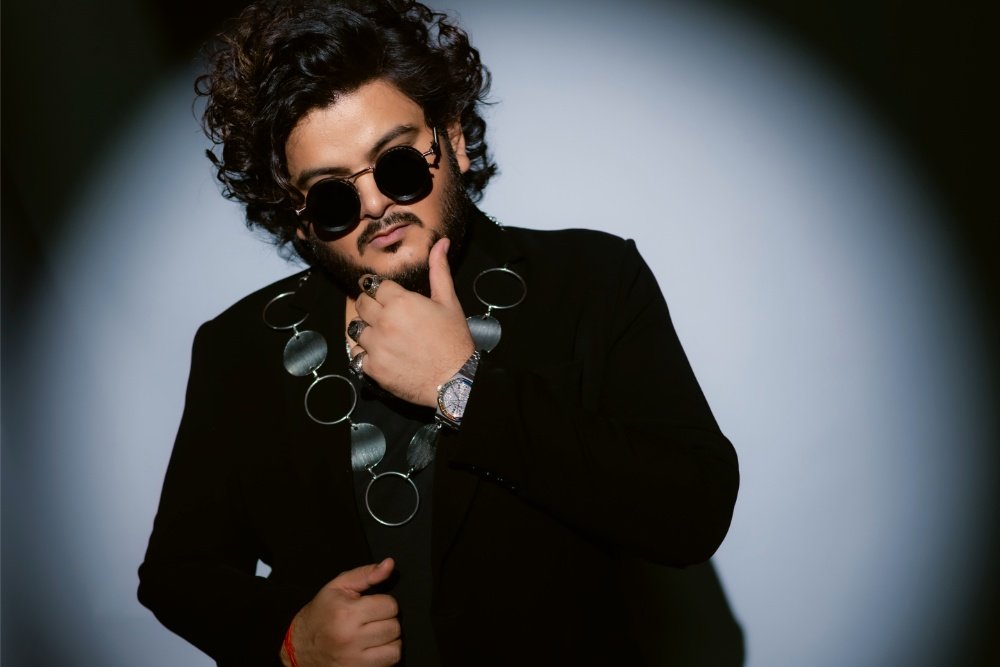 A bearded man with curly hair wears round dark sunglasses, a black jacket, and a large circular pendant necklace, posing thoughtfully against a lit backdrop.