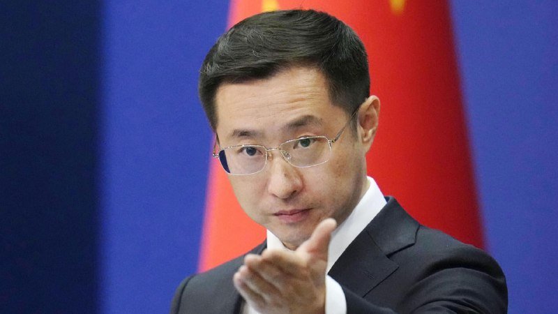 A bespectacled man in a dark suit gestures with an open hand in front of a red flag with yellow stars and a blue backdrop.