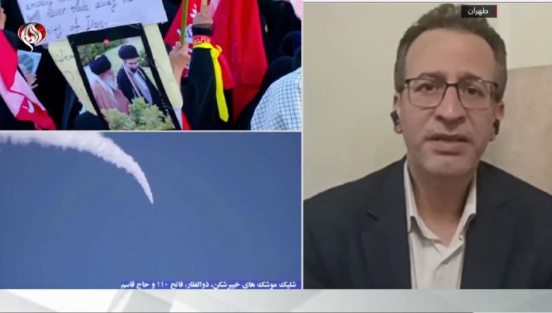 Split-screen news broadcast shows a bespectacled man in a suit on the right alongside footage of a crowd holding religious portraits, red flags, and a rocket's smoke trail over a body of water, with Arabic text overlay.