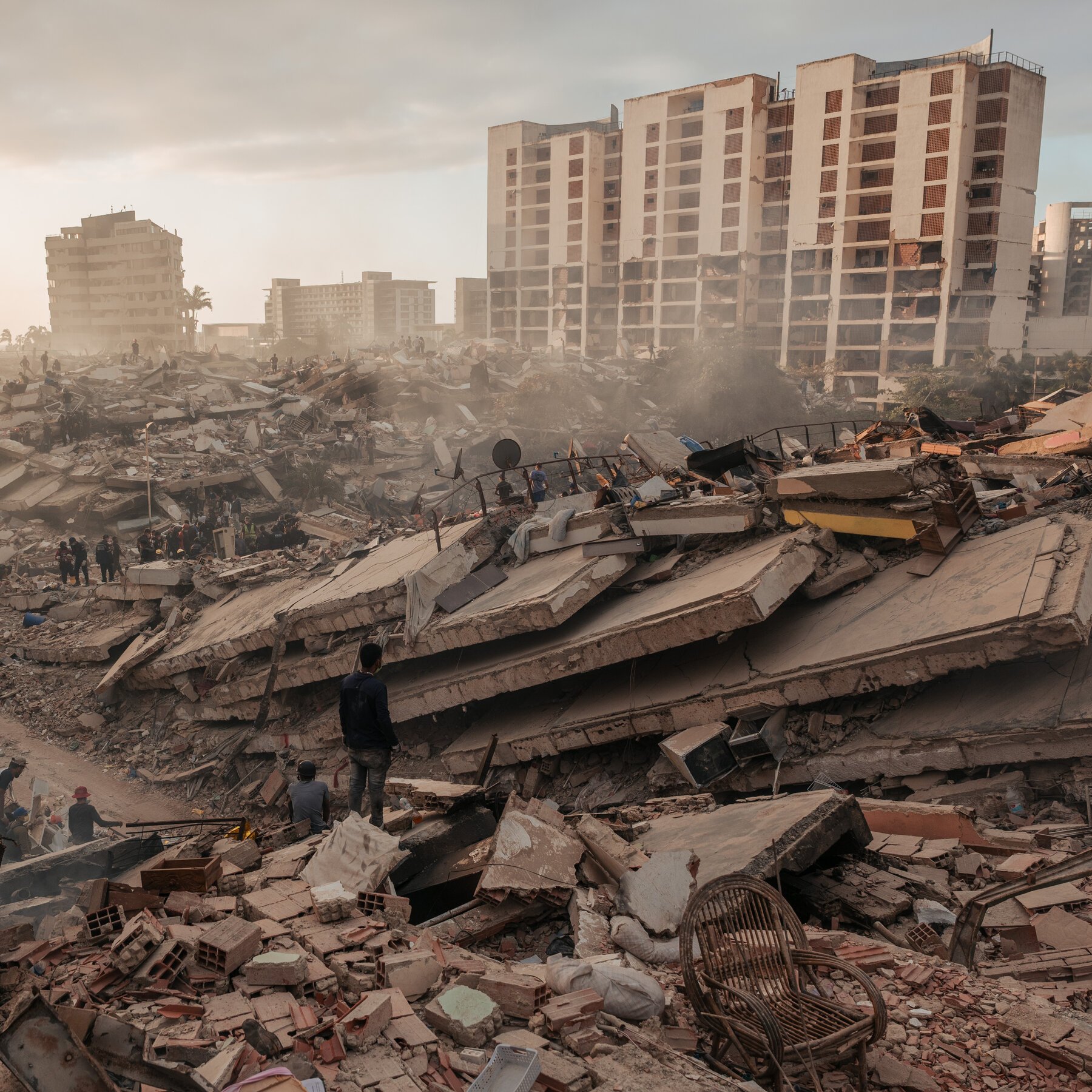 Rescue workers search through massive piles of concrete rubble and debris from a collapsed building, with damaged high-rise structures and dust visible in the background.