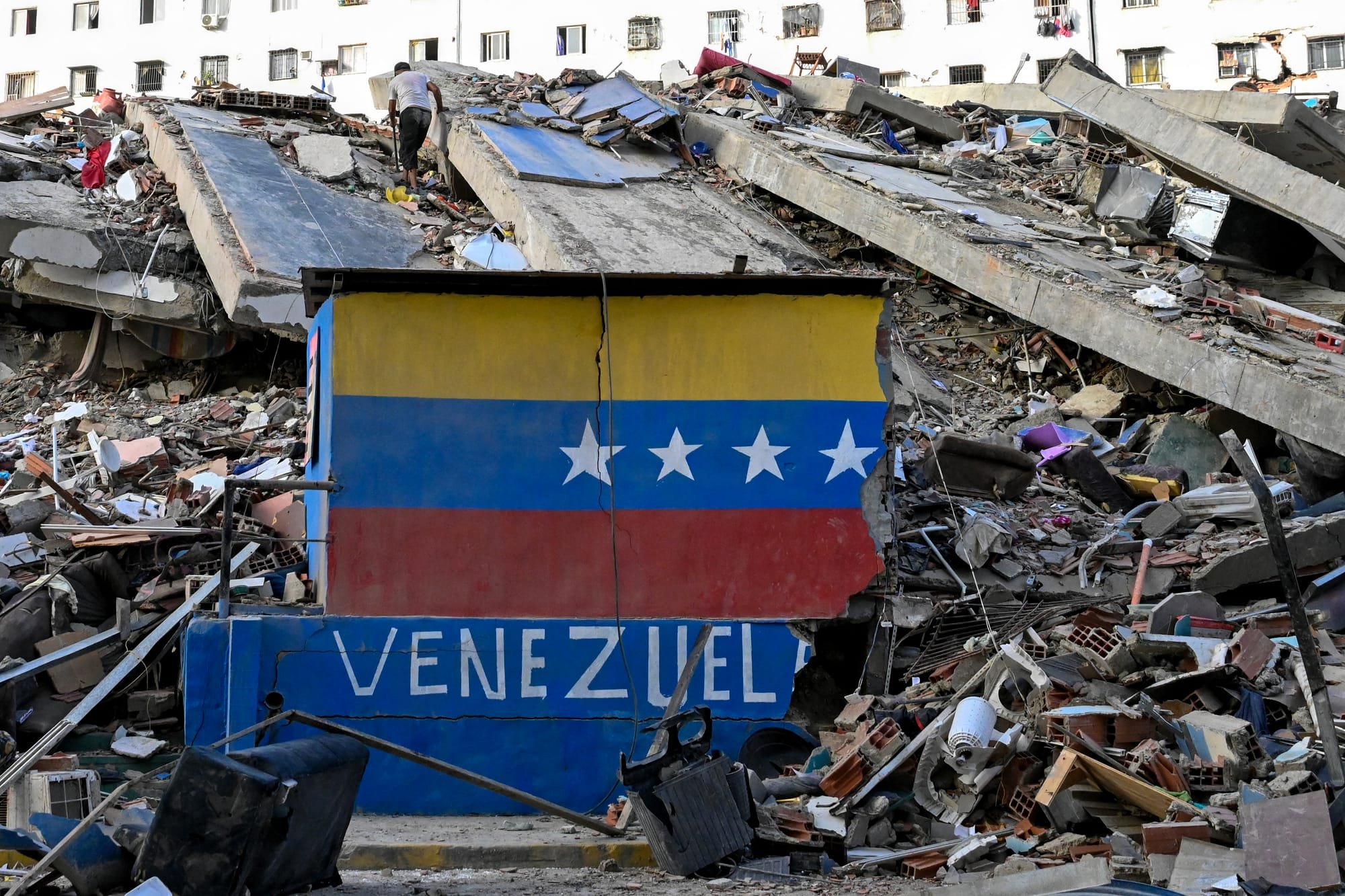 A damaged structure painted with the Venezuelan flag and the word "VENEZUELA" stands amid extensive rubble, with a person visible on the debris in the background.