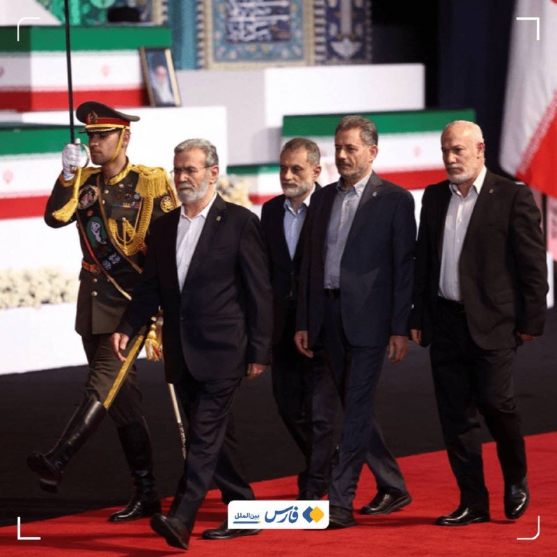 Five men walk in a line on a red carpet, led by a uniformed military officer carrying a sword, with an Iranian flag-draped platform in the background.