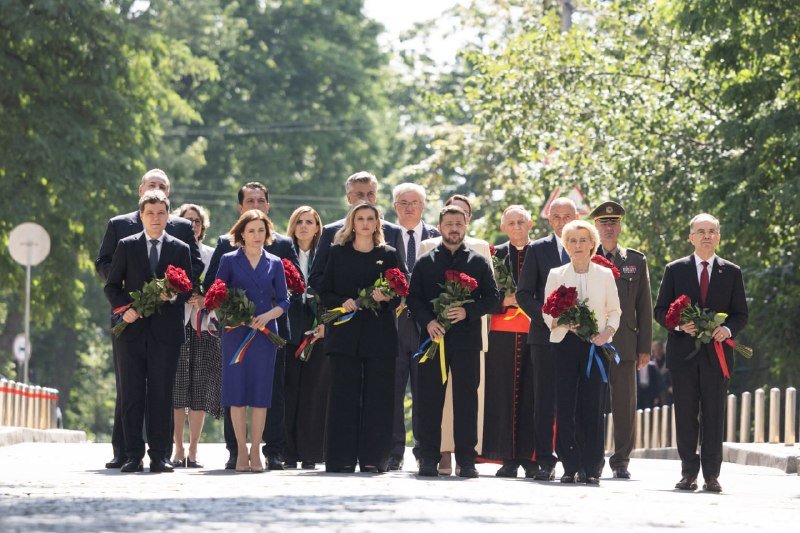 A group of formally dressed officials, including a man in a black shirt and a cleric in red robes, walk down a tree-lined street holding bouquets of red roses.