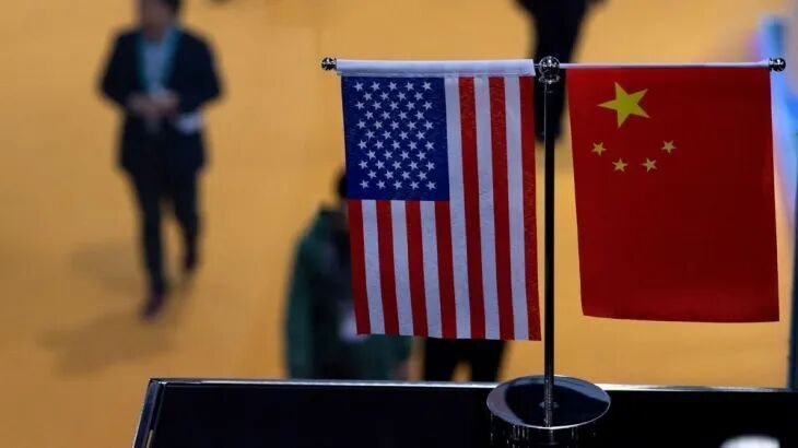 Small United States and Chinese flags stand side-by-side on a tabletop stand, with blurred figures of people walking in the background.