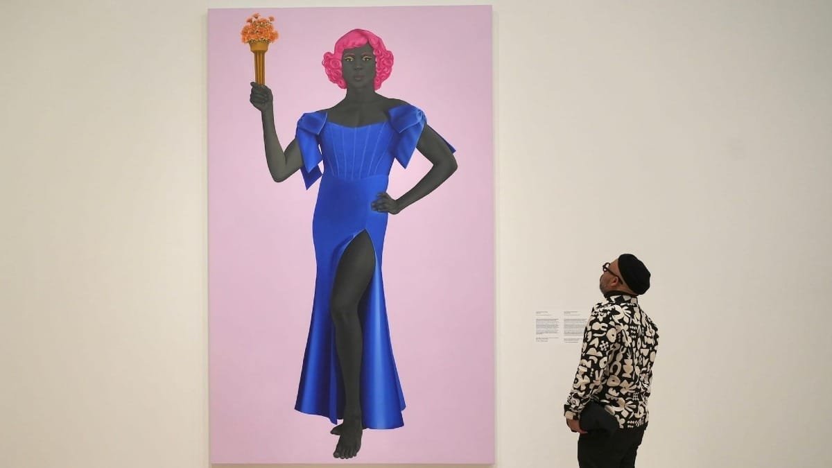 A man in a patterned shirt and beanie looks up at a large painting of a dark-skinned woman with pink hair in a blue gown holding orange flowers against a pink background.