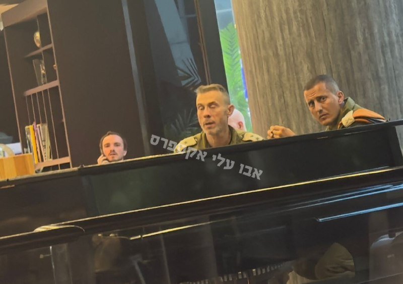 Three men in military uniforms sit behind a black grand piano indoors, with one speaking while another gestures, as Hebrew text appears overlaid on the image.
