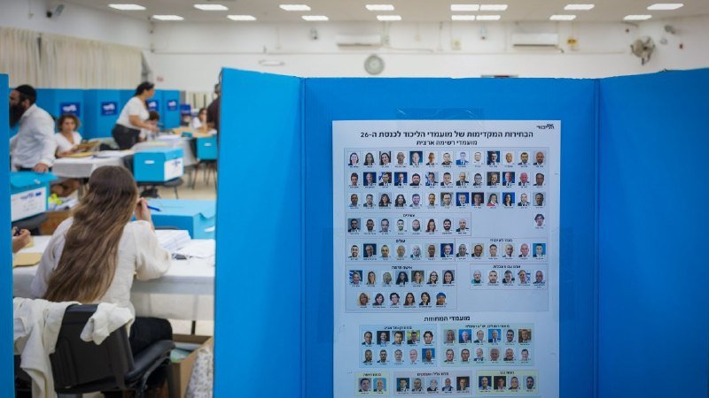 A woman sits at a desk in the background while a large blue display board in the foreground shows a Hebrew poster featuring rows of candidate headshots.