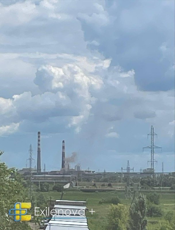 Thick black smoke and orange flames rise from an industrial facility with tall red-and-white striped smokestacks, viewed above a tree line.