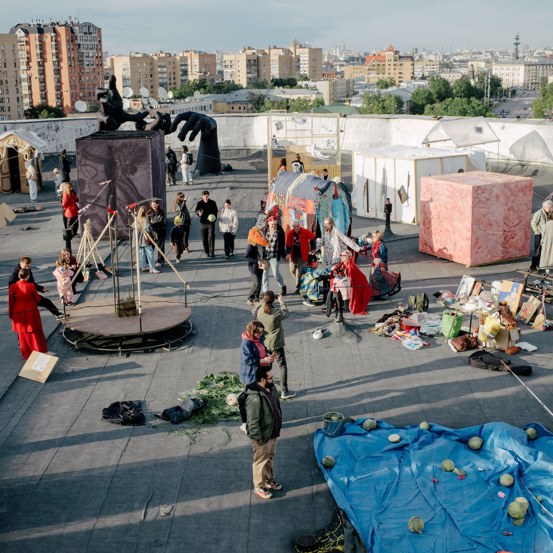 Visitors gather around large art installations, tents, and sculptural objects on a rooftop, with a cityscape visible in the background.