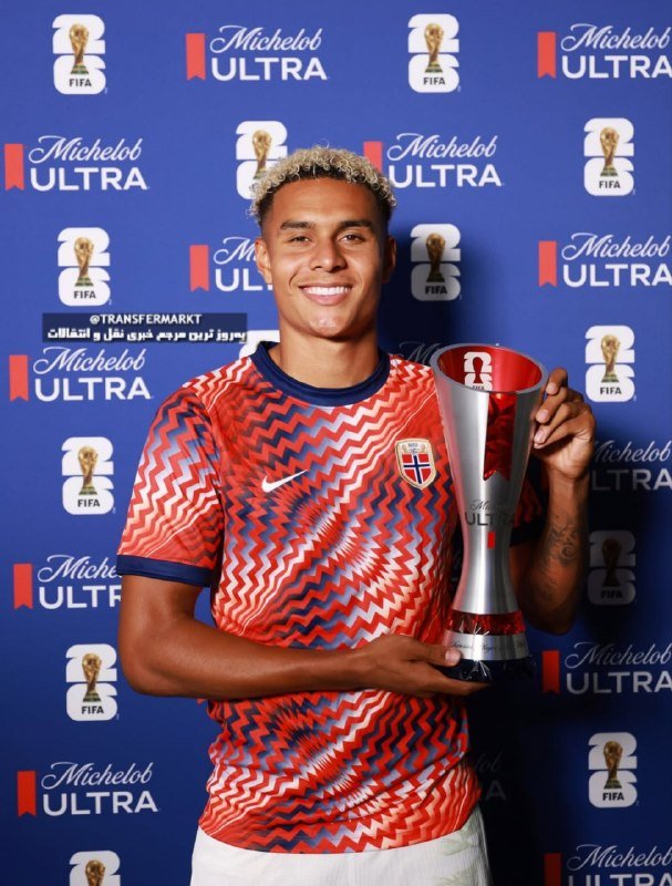 A smiling player in a red patterned Norway jersey holds a silver trophy against a blue backdrop featuring FIFA World Cup and Michelob Ultra logos.