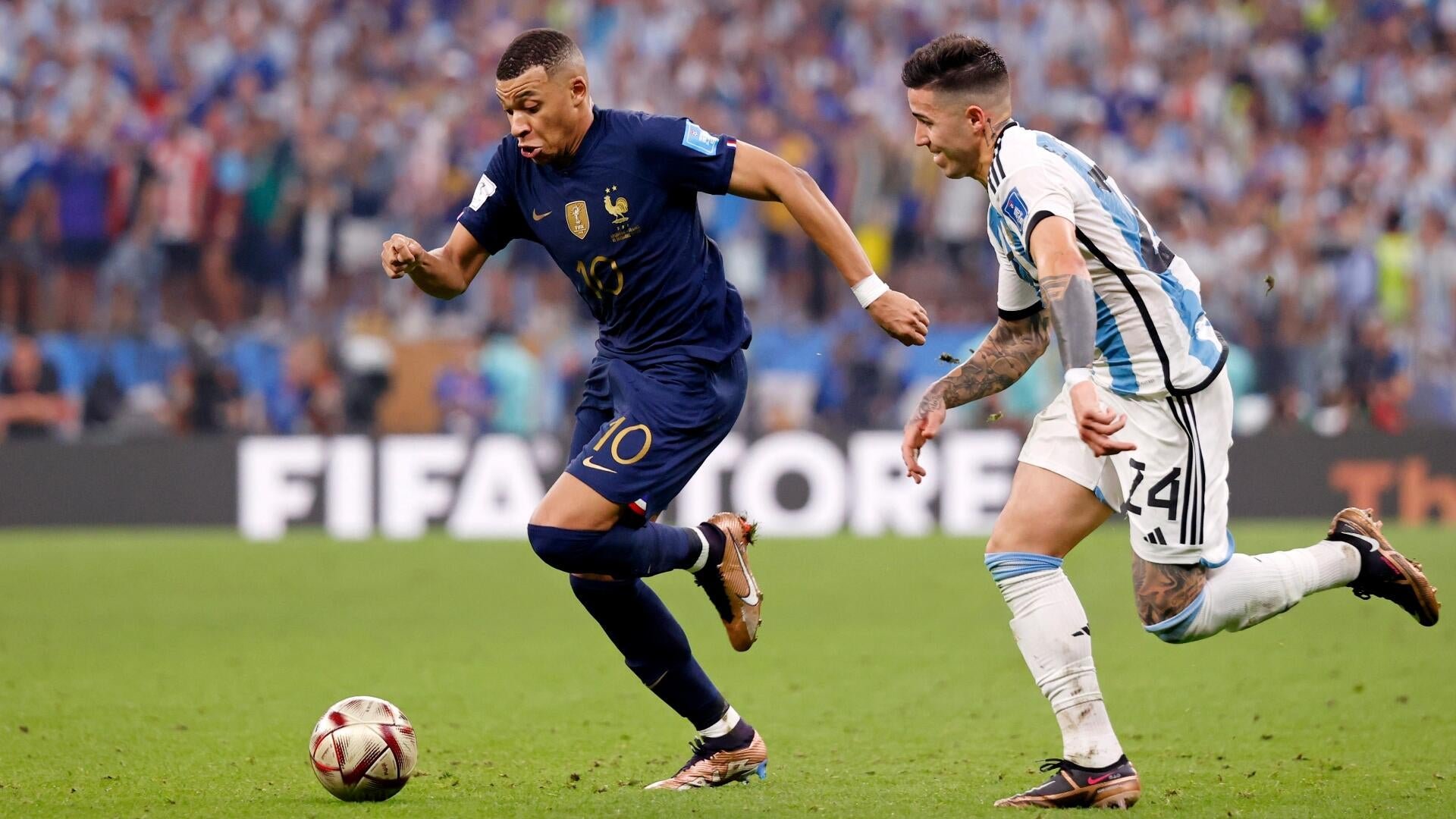 Two soccer players, one in a dark blue jersey numbered 10 and another in a white and light blue striped jersey numbered 24, sprint across a grass field pursuing a ball during a stadium match.