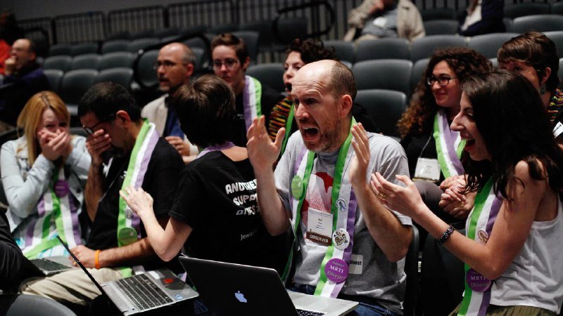 Audience members wearing green and white sashes react with open mouths and raised hands in a tiered auditorium, with laptops visible in the foreground.
