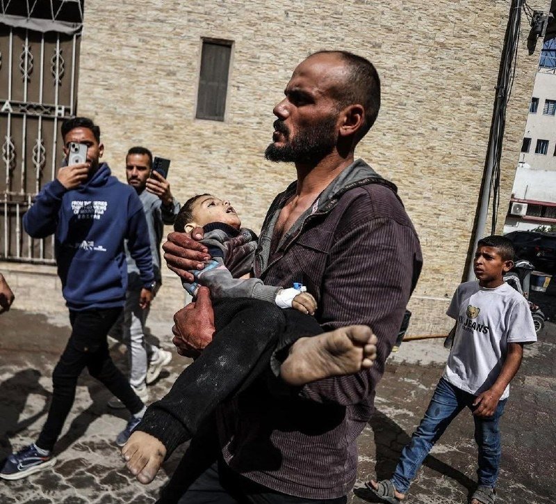A bearded man carries an injured child with a bandaged arm down a stone-paved street past onlookers holding up phones.