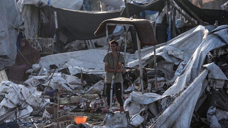 A young person stands among extensive debris and damaged tents in a destroyed encampment.