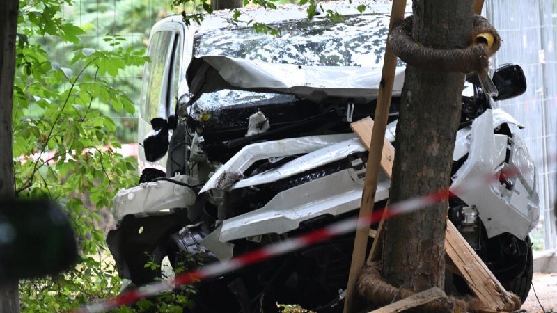 A severely damaged white vehicle with a crumpled front end sits beside a tree and red-and-white caution tape in a wooded area.