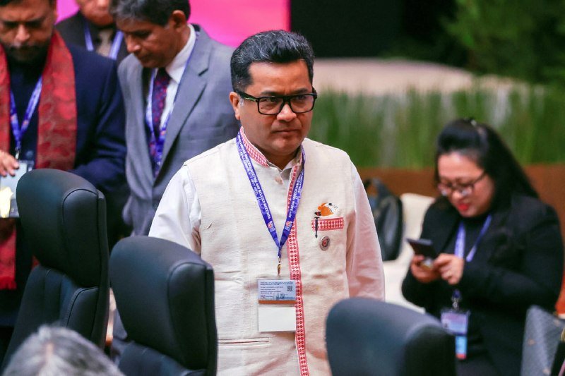 A bespectacled man wearing a lanyard, ID badge, and traditional embroidered vest stands among seated attendees at an indoor conference or meeting venue.