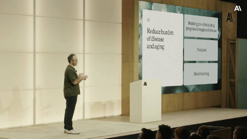 A man in a green shirt presents on stage beside a large screen displaying "Reduce burden of disease and aging" alongside an "AI" logo.