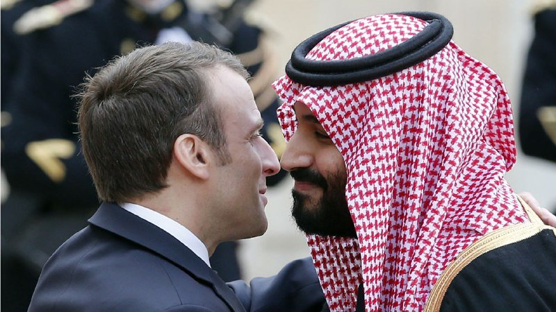 Two men, one in a dark suit and one in a red-and-white keffiyeh, greet each other by pressing their noses together, with uniformed figures blurred in the background.