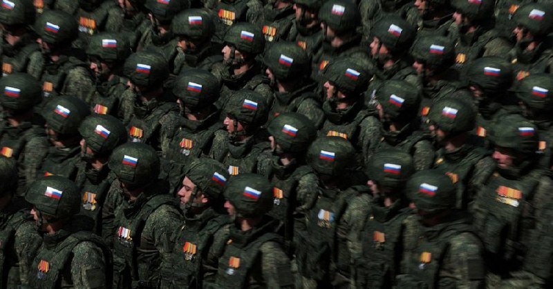 Soldiers in camouflage uniforms and green helmets bearing white, blue, and red flag patches march in tight formation, several wearing visible medals on their chests.