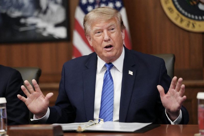 A blond man in a dark suit and blue tie gestures with both hands raised while speaking at a wooden table, with an American flag visible behind him.