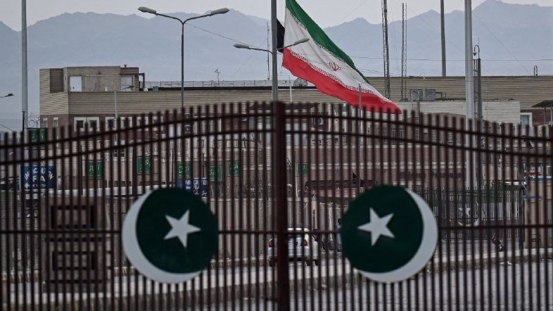 The Iranian flag flies behind a metal gate displaying Pakistan's green and white star symbols, with mountains and urban buildings visible in the background.