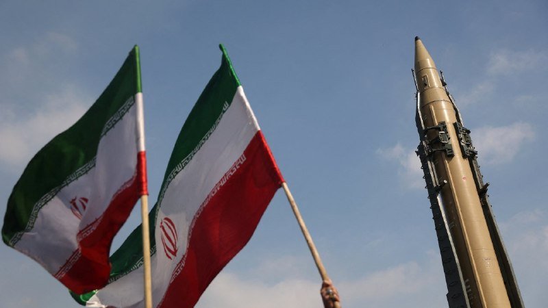 Two men in dark suits sit in chairs facing each other, holding documents, between a black flag with white calligraphy and the Iranian flag, with a framed photo on the table.