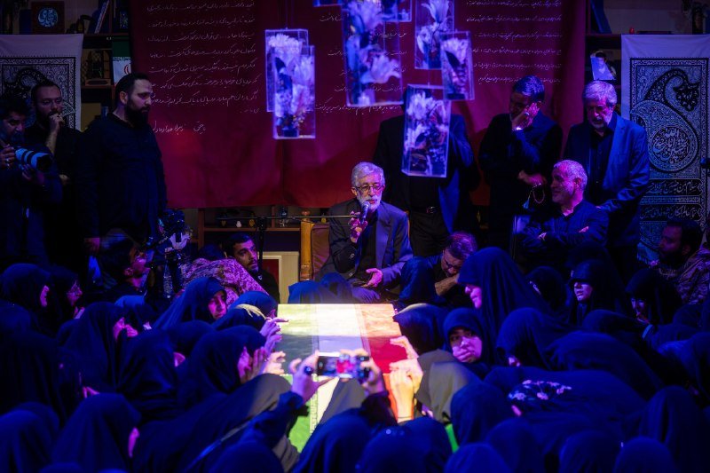 A man speaks into a microphone beside a flag-draped coffin, surrounded by women in dark headscarves taking photos with their phones in a dimly lit gathering.