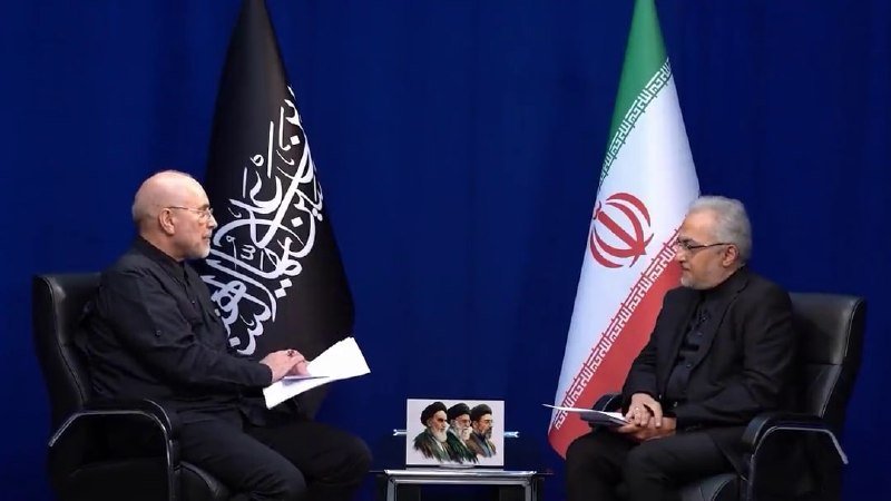Two men in dark suits sit facing each other in chairs, conversing between a black flag with white calligraphy and Iran's tricolor flag, with a small framed photo on a table between them.