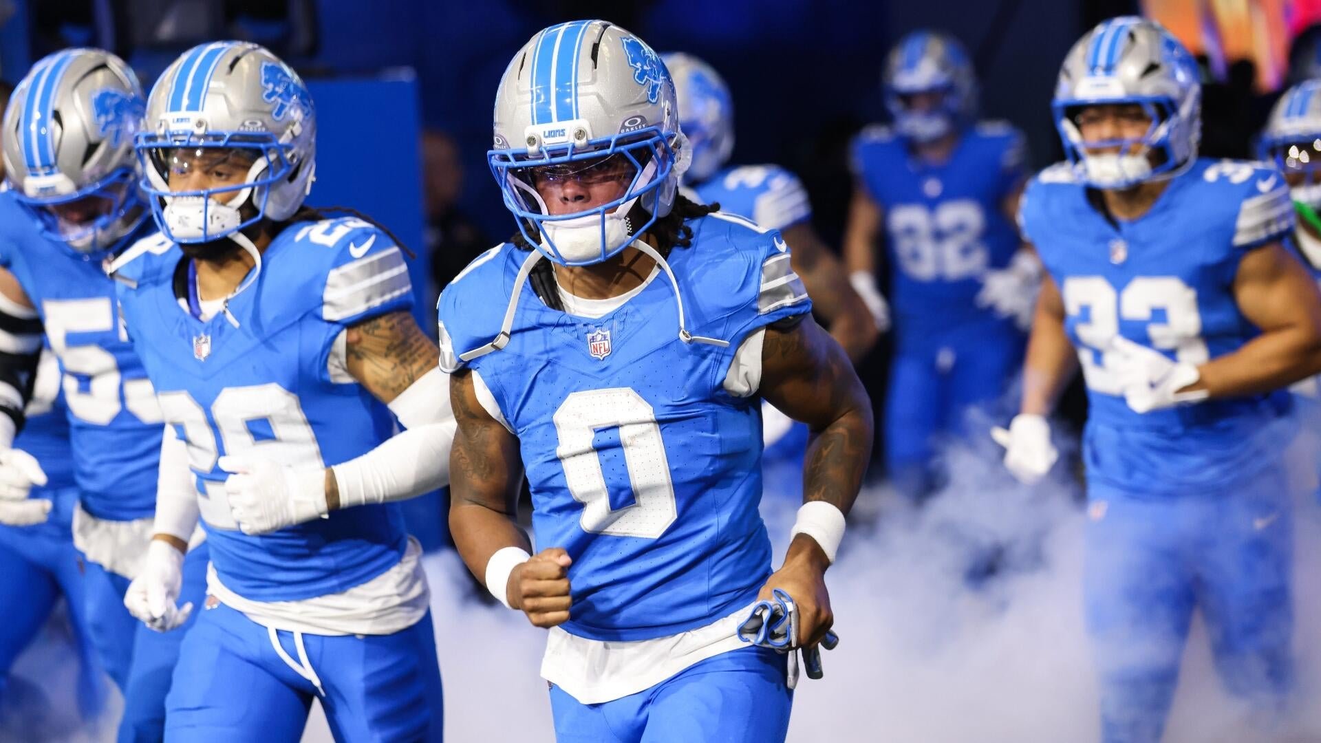 Football players in blue Detroit Lions uniforms and silver helmets run onto a field through smoke, led by a player wearing jersey number 0.