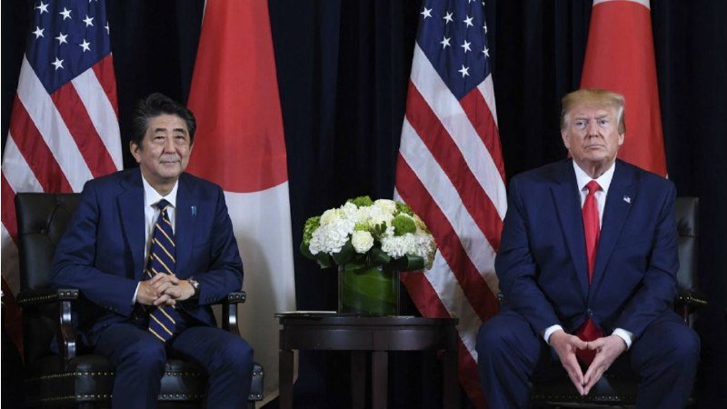 Two men in dark suits sit apart in dark leather chairs, hands clasped, with American and Japanese flags and a floral centerpiece between them.