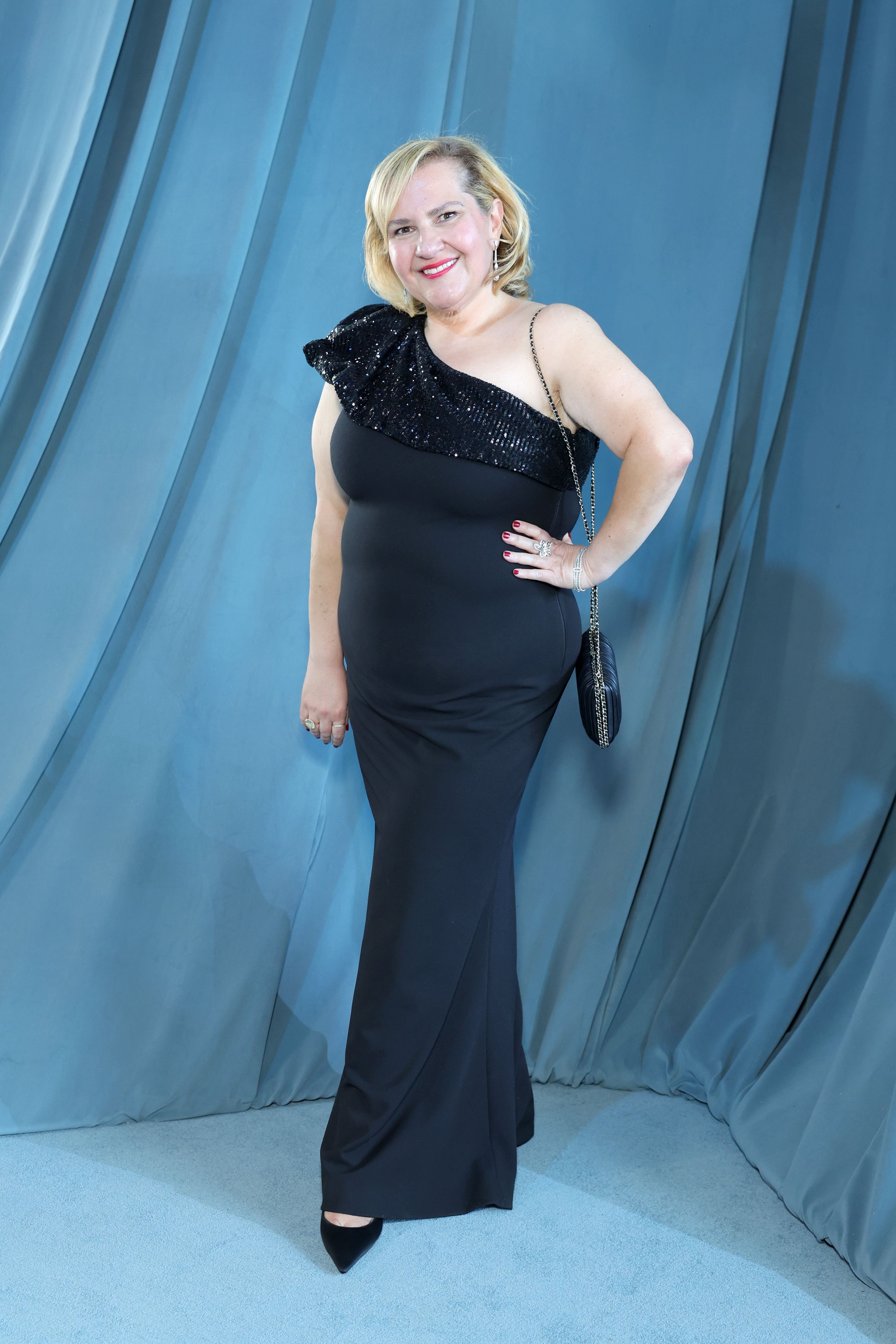 A smiling blonde woman in a black one-shoulder sequined gown poses on a carpet against a draped blue curtain backdrop.
