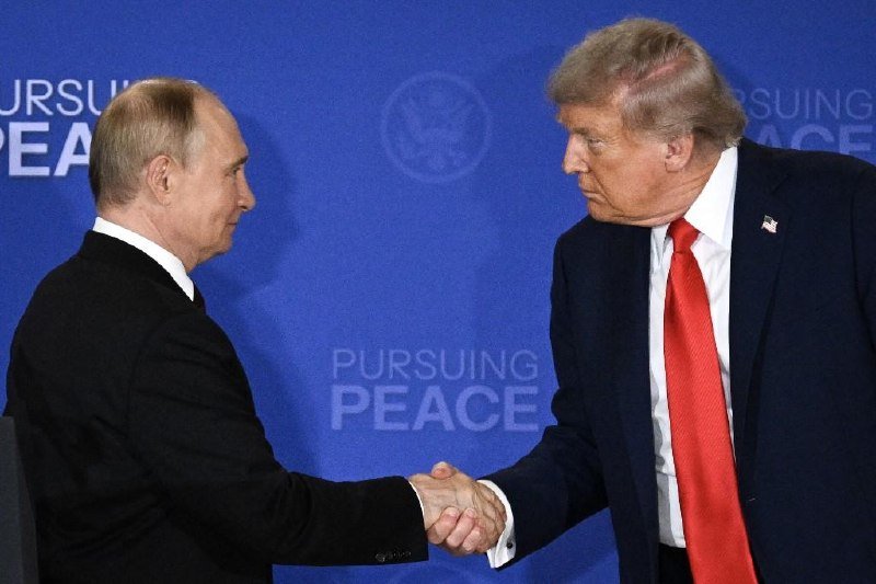 Two men in dark suits shake hands in front of a blue backdrop reading "PURSUING PEACE," with one wearing a red tie and an American flag pin.