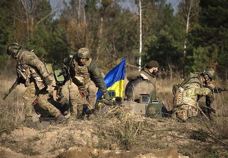 Armored soldiers in camouflage uniforms, one carrying a blue and yellow flag, move through tall dry grass near a forested area.