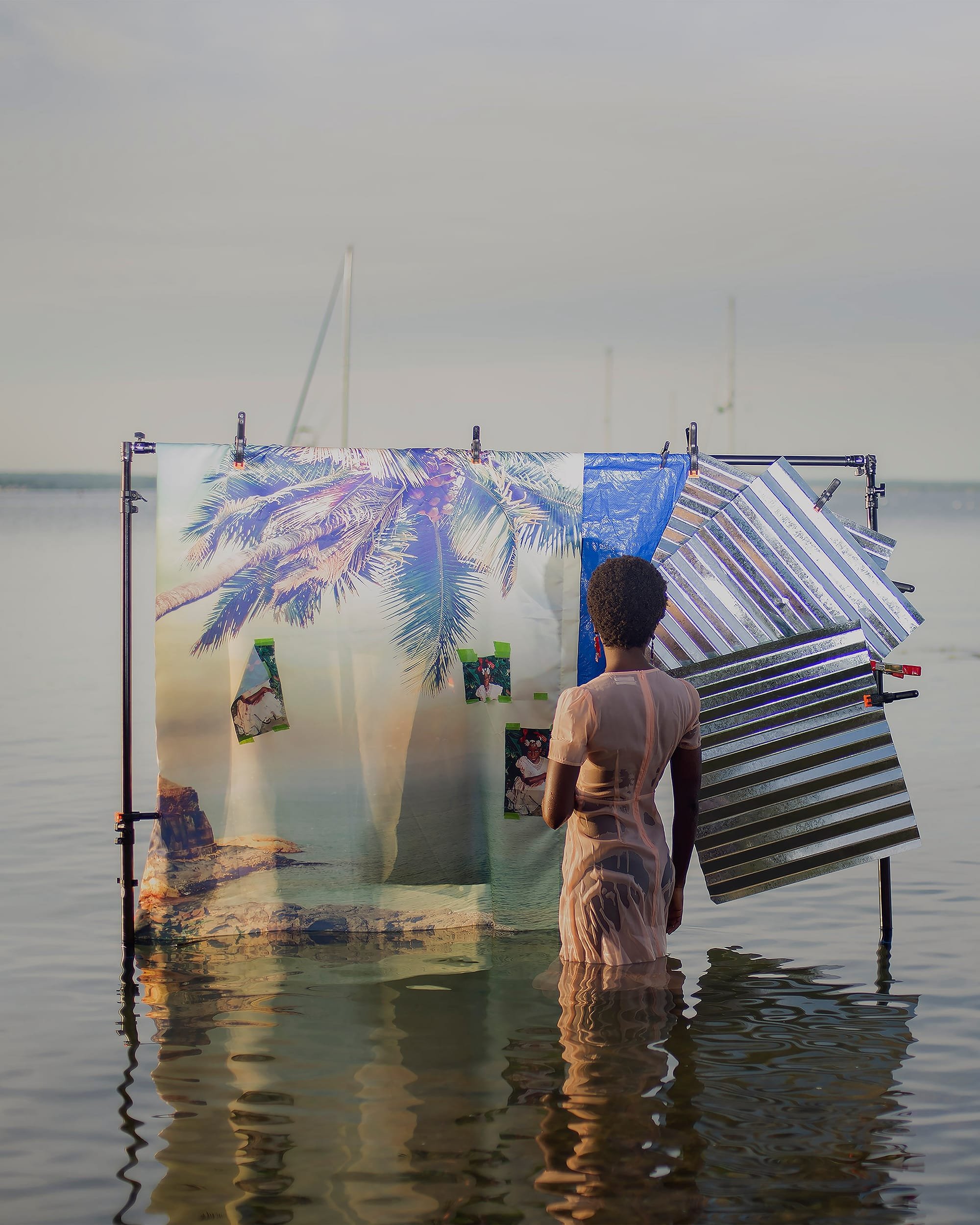 A person with short hair stands waist-deep in calm water, facing a metal frame holding a tropical beach backdrop and a silver reflector panel.