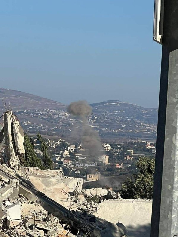 A plume of gray smoke rises above a hillside town viewed from a destroyed building's rubble, with mountains in the background under a clear sky.