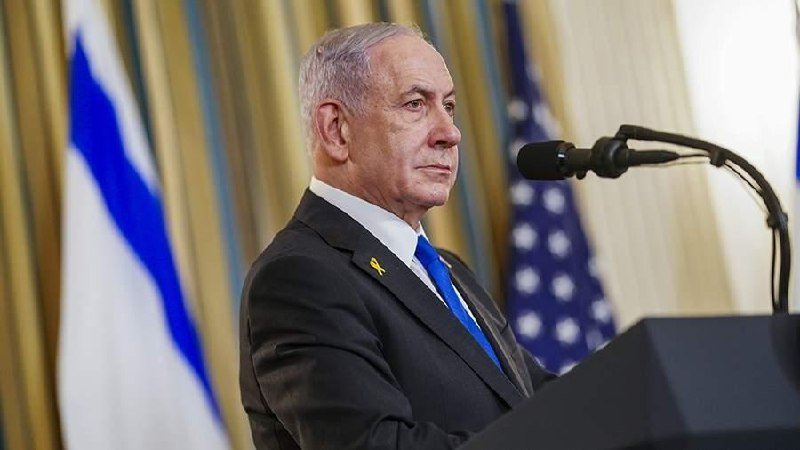 A gray-haired man in a dark suit and blue tie stands at a podium with microphones, a yellow ribbon pinned to his lapel, with Israeli and American flags visible behind him.