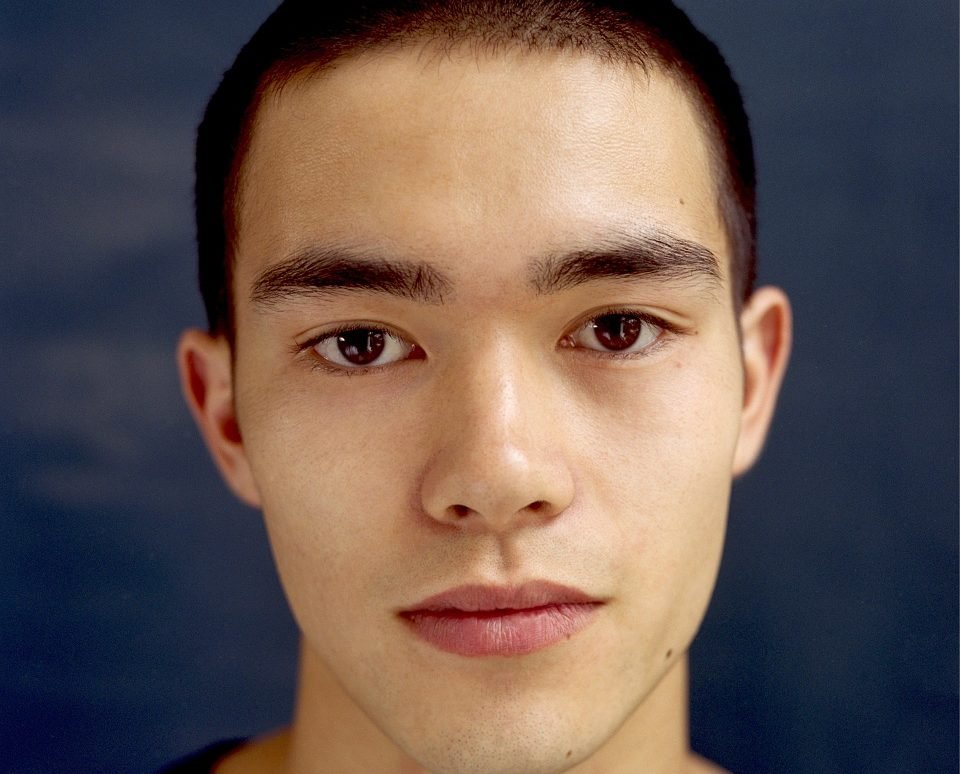 Close-up portrait of a young man with short dark hair looking directly at the camera against a dark blue background.
