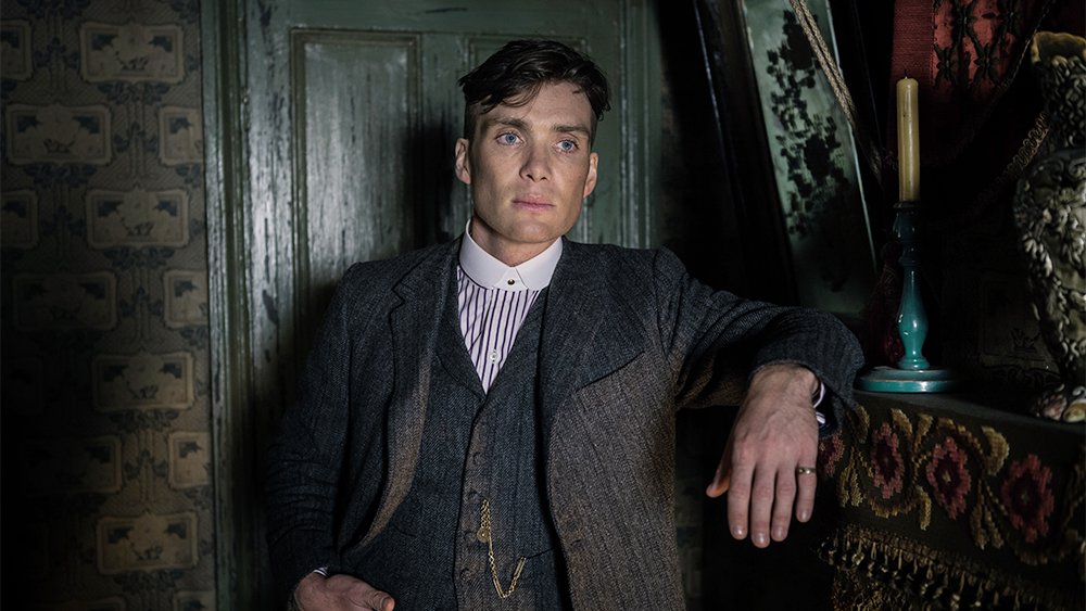A man in a gray tweed three-piece suit and striped shirt stands against a green paneled door, his arm resting near a lit candle on a tapestry-draped mantel.