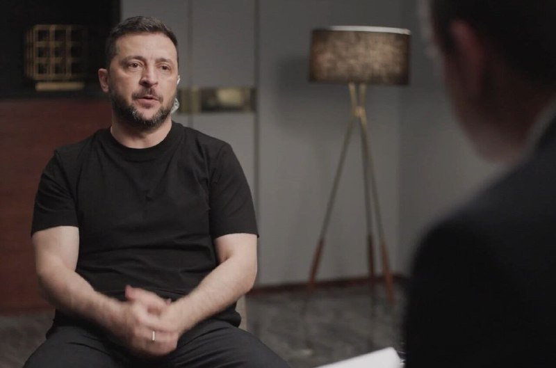 A bearded man in a black t-shirt sits with hands clasped, facing an out-of-focus interviewer holding papers, with a floor lamp in the background.