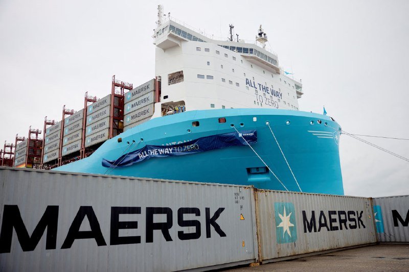 A massive blue and white Maersk cargo ship, bearing the slogan "ALL THE WAY TO ZERO," sits docked behind stacked shipping containers branded with the Maersk name and logo.