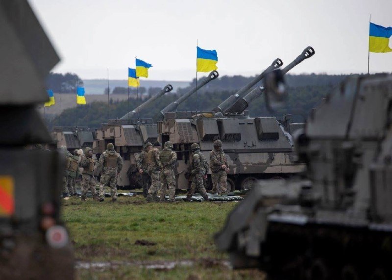 Soldiers in camouflage uniforms stand beside a row of self-propelled artillery vehicles mounted with Ukrainian flags in an open grassy field.